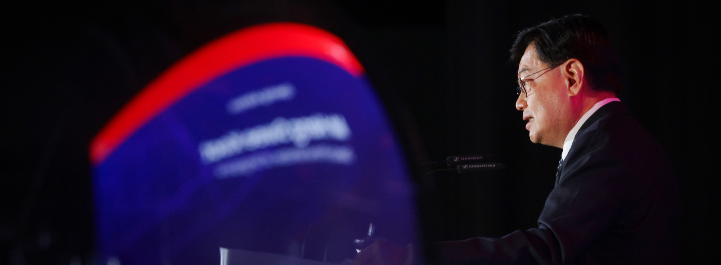 Man in suit speaking at podium with Sennheiser mics and blurred blue/red sign.
